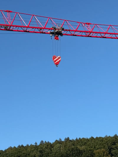 A red construction crane extends across a bright blue sky, its lattice boom reaching horizontally. Suspended from the crane's trolley is a red and white heart-shaped hook, intricately detailed with white stripes. Below the crane, a dense, green forest blankets a hillside. The image appears to have been taken in or near Bad Orb, Germany, likely at a construction site, given the presence of the crane.
