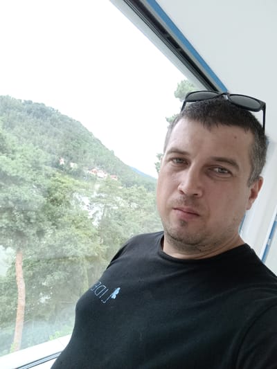 A man with short brown hair and a goatee is taking a selfie by a window. He is wearing a black t-shirt with a light blue logo on the left sleeve, which appears to be the word "TIDE" followed by a butterfly graphic. Sunglasses are perched on top of his head. Through the window, a green, tree-covered hillside with some buildings can be seen, along with a body of water. The sky is overcast and bright. This scene is located in Doksy, Czechia.