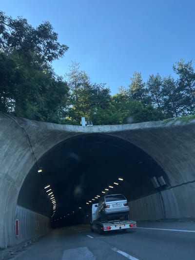 The image captures the entrance of a concrete tunnel under a clear blue sky, with lush green trees lining the top edge of the tunnel. A tow truck is visible inside the tunnel, carrying a silver car on its flatbed. The car has a license plate that reads "CO 5885 CX". The tow truck's license plate is "CO 2847 CT". The tunnel is illuminated by rows of lights along its ceiling. The location is identified as the city of Dolna Vasilitsa, Bulgaria.