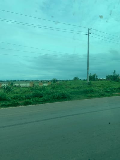 The image shows a roadside scene with a paved road in the foreground. Beyond the road, there is a grassy embankment topped with a line of vegetation and a low wall. A utility pole stands prominently on the embankment, with power lines extending across the sky. The sky is overcast and a greenish-blue hue, possibly due to atmospheric conditions or a filter. There are some dark spots on the surface of the image, which may be smudges or dirt on the lens. The general environment appears to be a rural or semi-rural area with signs of development, such as the wall and utility pole. It is difficult to ascertain the exact location within Laminga, Nigeria, based solely on this image.