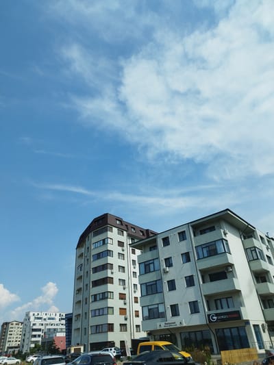 In Bragadiru, Romania, this image captures a street view dominated by modern apartment buildings and a clear blue sky with scattered white clouds. In the foreground, several cars are parked, including a black sedan and a yellow vehicle. The buildings are multi-story structures with contemporary architectural designs, featuring numerous windows and balconies. One building prominently displays signage for "GEORGE'S FISHERIES" and a "Restaurant" with Romanian text. The environment appears to be a developed urban area with a mix of residential and commercial spaces.