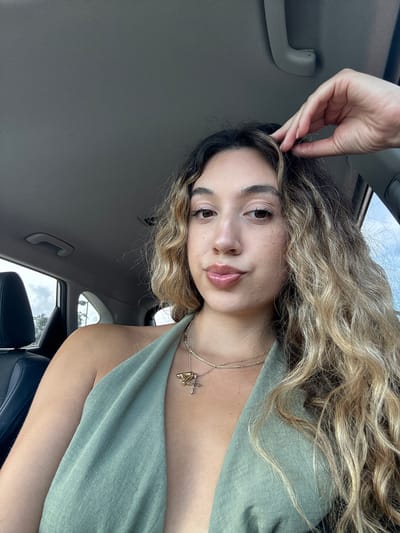 This is a vertical, close-up selfie of a young woman with wavy, blonde and brown hair, taken inside a car. She is looking directly at the camera, with a slight smile. Her right hand is raised, with her fingers gently touching the top of her head. She is wearing a green top and a necklace with a pendant. The background shows the interior of the car, including a gray handle on the ceiling and a portion of the car window, through which the sky and some trees are visible. The lighting suggests it is daytime, possibly late morning or early afternoon. The scene appears casual and relaxed.
