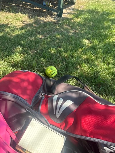An eye-level, close-up shot shows a red and gray softball bag with an open zipper, lying on green grass. A yellow softball rests on the grass to the left of the bag. Inside the bag, a spiral-bound notebook is visible. The background shows dappled sunlight filtering through leaves onto the grass and the edge of a dark structure. The scene appears to be outdoors in a park or backyard setting.