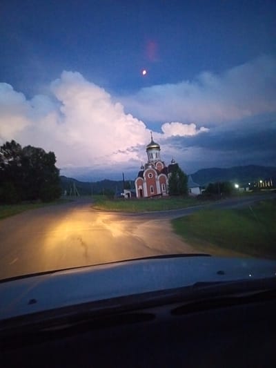 The image depicts a twilight scene in the city of Sadovyi, Russia. In the distance, a red and white church with a golden dome stands prominently. The church is flanked by trees on the left and a few houses and streetlights on the right. A dark road, illuminated by the headlights of an unseen vehicle, leads towards the church. The sky is a deep blue with dramatic clouds, and a faint, possibly red, celestial object is visible in the upper portion of the frame. The foreground shows the interior of a vehicle, with the dashboard and windshield visible.