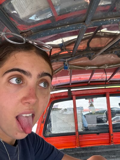A young female is photographed in Tambon Tha Uthae, Thailand, occupying the rear passenger area of a red utility vehicle, likely a modified pickup truck or songthaew. Her fair-skinned face is partially visible in the foreground, angled upwards and to the right, with prominent dark eyebrows and green eyes looking sideways. Clear-framed glasses rest on her forehead. Her mouth is open, and her tongue is extended. She wears a dark t-shirt and a silver chain necklace.
The vehicle's interior features a red metal frame structure and an orange-red tarp canopy secured by blue ropes overhead. Behind the subject, a red-painted metal partition and a rear window are visible. Through this window, a light-colored document or chart is taped, and reflections reveal part of a driver's cabin, including a steering wheel and a hanging white air freshener. Exterior elements visible through the window suggest another vehicle and an outdoor environment, possibly a road. The lighting indicates daylight conditions.