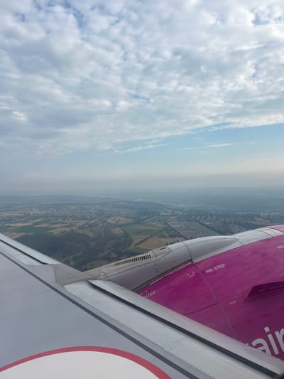 This image, taken from an airplane window, provides a view of Echterdingen, Germany, from an aerial perspective. The foreground is dominated by the wing of a Wizz Air airplane, characterized by its distinctive pink engine housing and white wing. The engine casing is marked with safety warnings such as "NO STEP" and "NO GRAB." Below the wing, the landscape of Echterdingen unfolds, revealing a patchwork of agricultural fields, forested areas, and the urban sprawl of the city. Buildings and infrastructure are visible, interspersed with green spaces. The sky above is filled with a mixture of white and grey clouds, with patches of blue sky peeking through, indicating a partly cloudy day. The overall impression is of an aerial view of a European city on a day with mixed weather conditions.
