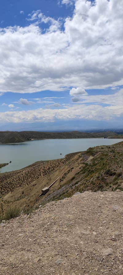 The image shows a large body of water, possibly a reservoir or lake, with a light greenish-grey hue, surrounded by hilly terrain. The foreground consists of a dirt and gravel slope. A concrete structure with a pipe extending down the hillside toward the water's edge is visible. The landscape appears arid with sparse vegetation. The sky is partially cloudy with patches of blue sky visible above. In the distance, there are mountains. The location of the image, Lanjazat, Armenia, is not readily apparent within this shot. There are no people, clear subjects or overt actions depicted. The scene is a landscape photograph of a reservoir and its surrounding area.