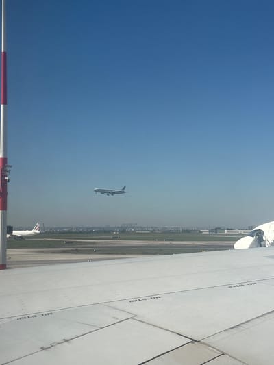 This daytime image, taken from what appears to be an airplane window or an elevated airport structure, depicts a bustling airport scene under a clear blue sky, with a hazy horizon line in the distance.
In the foreground, the clean white surface of a large airplane wing occupies the right side of the frame. Multiple "NO STEP" warnings are stenciled clearly on the wing. At the bottom of the image, a red and white striped barrier or pole is partially visible.
A prominent subject in the mid-ground is another large airplane, airborne against the blue sky, suggesting it is either taking off or landing. Its underside is visible as it ascends or descends. Further towards the lower center of the image, the white tail fin of a third airplane can be seen parked on the tarmac.
The background features an extensive airport environment with runways and taxiways, stretching towards a distant urban or industrial landscape with buildings and structures, somewhat obscured by the haze. The clear weather and visible aircraft activity indicate a busy operational day at the airport. This scene is consistent with the location context of Tremblay-en-France, home to a major international airport.