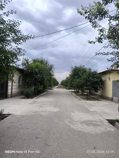 The image depicts a street in Pakhtakor, Uzbekistan. The street is lined with single-story residential buildings on either side, separated by trees and vegetation. The street surface is paved with a light-colored material, possibly gravel or asphalt. The sky is overcast with gray clouds. Electrical wires are strung overhead. No people are visible in the image. In the distance, a pile of gravel or construction material is present. The photograph was taken with a Redmi Note 13 Pro+ 5G, with the date and time stamp indicating 27.05.2026 12:19.