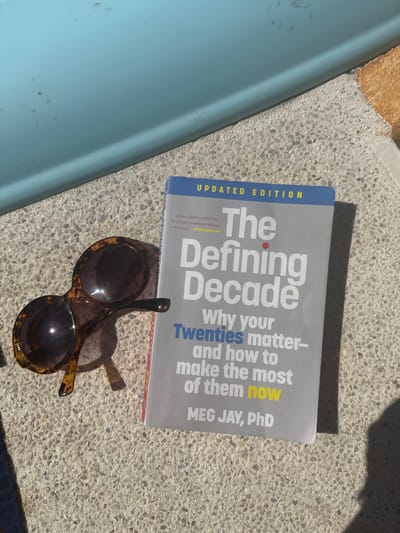 The image shows a book titled "The Defining Decade: Why your Twenties matter- and how to make the most of them now" by Meg Jay, PhD. The book is an updated edition and features a quote, "A four-alarm call for the 50 million twentysomethings in America" -Kirkus Reviews. A pair of brown-spotted sunglasses is placed on the book, casting a shadow. The items are resting on a rough, light-colored surface. A blue object is visible in the top left of the frame.