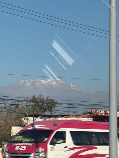 The image displays an urban landscape in Santa María Zacatepec, Mexico, viewed through a reflective surface that exhibits diagonal light streaks. The upper portion shows a clear, light blue sky. Below the sky, a distant range of snow-capped mountains is visible. Numerous dark power lines horizontally traverse the mid-ground. Beneath the power lines are sparse, bare-branched trees and some green foliage. Portions of urban structures are visible behind the trees and lines, including brickwork and a partially obscured, weathered sign reading "MADERERIA" on the right. A grey utility pole stands vertically on the far right side of the frame. In the foreground, a white and bright pink (magenta) passenger van is in motion from left to right, indicated by slight blurring. The front of the van displays "ZACATEPEC," "R-25," and "U-78-." Faint reflections of individuals are discernable within the van's windows.