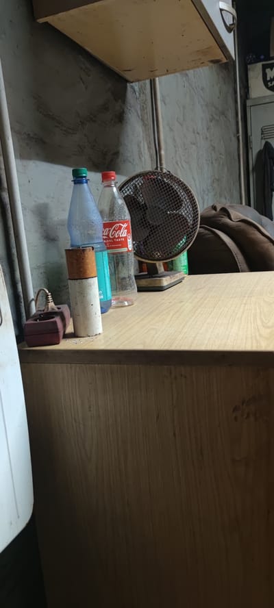 The image depicts a close-up view of a wooden surface, likely a desk or cabinet, in what appears to be a storage area or workshop. On the surface, there are several objects arranged. To the left, a red electrical outlet with a plug and cord is visible. Next to it stands a white cylindrical object with a brown top, possibly a ceramic pot or a container. Behind these, two plastic bottles are placed: one is clear blue with a green cap, and the other is clear with a red cap and a Coca-Cola label. Further to the right, a small, vintage-style fan with a metal grille is positioned. The background consists of a textured, grayish wall and a hint of a brown upholstered object, possibly a couch or chair, to the far right. The overall lighting suggests an indoor setting, possibly with artificial light. The scene is devoid of any human presence or activity. The location is not specified in the image or its provided context.