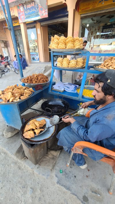 In an outdoor setting in Unknown, Pakistan, a man with a beard, wearing a blue outfit and black cap, is seated on an orange chair and frying food in a large wok. He is using a slotted spoon to transfer fried items from the wok to a metal bowl. The cooking area is part of a food stall, which features multiple tiers holding various fried foods, including what appear to be samosas and other snacks. The stall is blue and has stacks of flatbread or similar items on the upper shelves. A newspaper is placed on one of the shelves. A row of shops is visible behind the stall, with signage in a local language. Motorbikes are parked nearby, and other people can be seen seated or walking in the background. The ground is concrete or pavement.