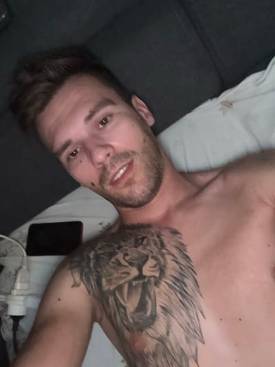 The image is a close-up, high-angle selfie of a shirtless man lying on a bed. The man has short, brown hair, brown eyes, and stubble. He is smiling faintly. On his chest and left shoulder, he has a large tattoo of a lion's head. To the left of the man, a smartphone and a white electrical plug are visible on the bed. The background consists of a dark gray surface, likely a headboard or wall, and a white pillow. There are no discernible elements in the image that would indicate its location in Sesto Calende, Italy.