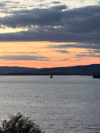 The image depicts a wide body of water, likely a river or lake, with a distant, tree-lined shore and rolling hills in the background. A small sailboat is visible in the middle distance, its sail partially furled. The sky above is a blend of dramatic clouds, with dark, stormy-looking clouds at the top and a band of warm orange and yellow hues near the horizon, suggesting either sunrise or sunset. In the foreground, the dark silhouette of a tree with dense foliage is visible on the left side, partially obscuring the water. The overall scene conveys a sense of calm and natural beauty. The location is identified as the Golubac Municipality in Serbia.