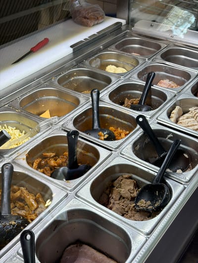This image depicts a commercial food display counter in ตำบล ไม้ขาว, Thailand. The foreground is dominated by a grid of stainless steel food pans, commonly used in buffet or sandwich bar setups.
From left to right and top to bottom:
* The top-left area behind a clear glass partition features a white cutting board with a red-handled utility knife resting on it. A plastic-wrapped food item is partially visible behind the glass.
* The upper row of food pans (partially visible) contains a creamy yellow spread, and sliced pink processed meat.
* The second row includes a pan filled with shredded yellow cheese sticks, an empty pan, a pan with an orange-colored stew containing diced items (possibly vegetables or fruit), and a pan with a dark brown, chunky mixture.
* The third row contains a pan of light-colored meat pieces in a brown sauce, an empty pan, a pan with grilled chicken strips, and a pan with a brownish, textured paste or spread.
* The bottom row (partially visible) shows a large slab of dark pink meat in a pan.
* Numerous black plastic serving spoons are present in many of the filled food pans.
* The stainless steel surfaces exhibit minor condensation or moisture. No individuals are visible in the scene. The overall environment suggests a clean, prepared food service area.