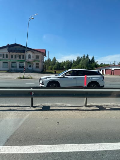 In Shobashkarkasy, Russia, on a sunny day, a white SUV is observed driving on a highway. The vehicle is in motion, captured from the side, with its windows tinted. A red reflective marker is attached to a pole beside the vehicle on the highway. In the background, a two-story building with a red roof houses a store and a cafe. The store has signage that reads "ВАШ МАГАЗИН ПРОДУКТОВ" (Your grocery store) and the cafe is marked as "КАФЕ". Further in the background, a line of evergreen trees stands against a clear blue sky. A red and white building with garage doors is also visible. The foreground features a gray highway with a metal guardrail.