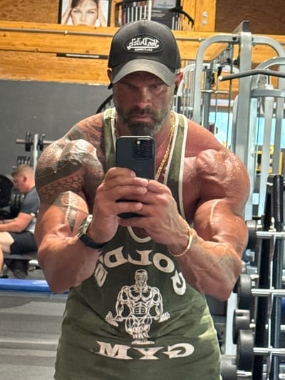 A muscular man is taking a selfie in a gym, seemingly in Jemelle, Belgium based on his "Gym de Jemelle" shirt. He is wearing a black baseball cap, a gold chain and bracelet, and a black watch. He has extensive tattoos on his arms. The gym equipment, including weights, is visible in the background, and another person is working out in the mirror's reflection. There is also a poster in the background.