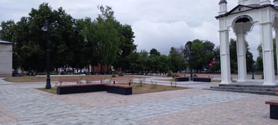 The image depicts a paved public square in the city of Kirsanov, Russia, featuring a white architectural structure with arched openings and columns, likely a gazebo or monument. Several park benches with wooden slats and dark bases are strategically placed throughout the square, some in L-shaped configurations. A lamppost with a decorative base and a single light fixture stands near the center-left of the frame. Lush green trees, including a tall birch tree, line the background, with a brick building visible on the far left. The sky is overcast with heavy clouds. The ground is paved with interlocking stones in varying shades of grey and tan, with patches of dry grass and small shrubs surrounding the benches. The scene appears to be unoccupied by people.