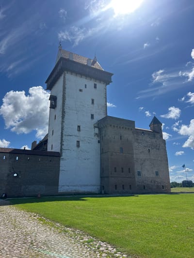 This is a vertical, eye-level, outdoor, daytime shot of the Narva Castle in Narva, Estonia. The castle is a large, stone structure with a white-plastered tower and a darker, brick-like base. It features several windows of various sizes and a dark, pitched roof on the tower. To the right of the castle is a well-maintained, bright green lawn. In the foreground, a cobblestone path leads alongside the grass. The sky is a vibrant blue with scattered white clouds, some of which are wispy and streaky, indicating a bright and sunny day with a gentle breeze. The sun is visible in the upper left corner of the image, casting a bright glare and lens flare. A flag is visible in the distance flying from a pole on top of a hill behind the castle. The overall mood is peaceful and serene, showcasing a historical landmark on a beautiful day.