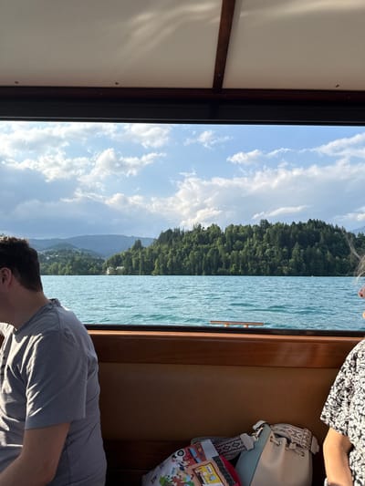 The image shows the interior of a boat on Lake Bled, Slovenia, taken from the perspective of a passenger looking out the window.  The coordinates provided correspond to a location on the lake near the small island with the church.  A man wearing a gray shirt sits on the left side of the frame.  A woman is seated on the right.  She is wearing a black and white patterned dress and is out of focus.  The woman has a white purse on the bench next to her.  The man and woman are looking forward.  There is a blue and red book and a colorful pillow on the bench in front of the woman.  The window frames a view of Lake Bled with the forested island and church in the distance.  The water is blue and ripples gently.  A mountain range is visible in the background.  The sky is blue with white fluffy clouds.