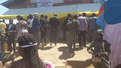 The image shows a gathering of people in front of a building in Kirumba, Democratic Republic of Congo. The building appears to be a government office, as indicated by the sign that reads "BAHATU MUSANGA DU NORD-KIVU ERASION UN LEADER QUI ECOUTE SA POPULATION." The people are mostly men, some wearing military uniforms. A soldier in camouflage fatigues stands guard near a blue and white flag, and a white tent is visible in the background. There are several people in the foreground, some standing and others sitting on the ground. The ground is uneven and dusty, and there is a ditch to the right of the image. The image appears to have been taken during the day.