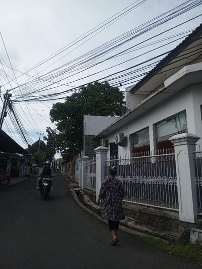 A residential street scene presents an overcast sky tangled with overhead wires. A woman walks away from the viewer on the right side of the road, adjacent to a white fenced property. A motorbike with two riders approaches in the distance. The street is dark asphalt, flanked by sidewalks and fences. Buildings are visible, including a white structure with dark-framed windows. A large tree adds a touch of greenery to the scene.