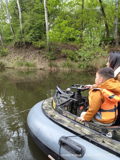 In Chlumec nad Cidlinou, Czechia, a small boat navigates a waterway flanked by a grassy bank topped with trees. A young boy wearing an orange life vest sits at the boat's helm, positioned near a black steering wheel. An adult female is partially visible beside the child. The boat itself has a gray inflatable border and a wooden-patterned interior. The water is dark and reflects the surrounding foliage.