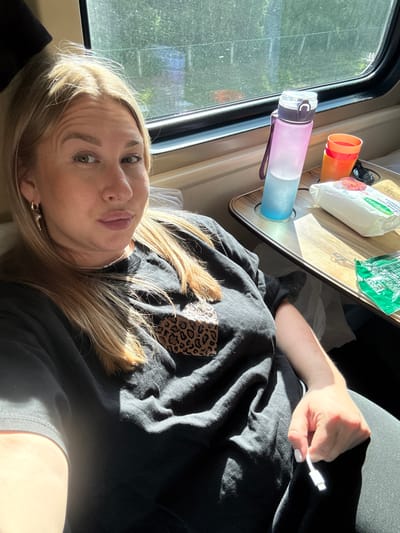 The image depicts a blonde-haired woman resting inside what appears to be a vehicle, likely a camper van, in Okulovka, Russia. She is lying down, looking towards the camera with a slight pout, illuminated by bright sunlight streaming through a window. She wears a black long-sleeved top with a leopard print patch on the upper arm. Her left hand, featuring white nail polish, is visible holding a white charging cable.

The interior of the vehicle includes a light-colored window frame and a wooden-patterned shelf or table. On this surface, a two-toned pink and blue water bottle, an orange cup, and sunglasses are placed. Two packages are also present: a white one bearing the text "Calypso" and "OSO," and a green one with "1997" partially visible.

Outside the window, blurred green foliage suggests a natural, possibly wooded, environment. The strong sunlight indicates daytime, casting pronounced highlights and shadows, particularly on the woman's hair and the objects on the shelf. The scene captures a moment of rest or leisure during a journey or stop.