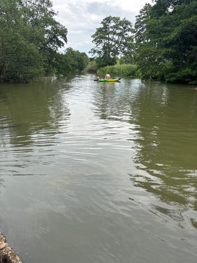 A narrow river flows between trees on either bank under a cloudy sky. The water is calm, reflecting the sky. A small yellow kayak with two people is visible in the distance, with one person paddling. The scene appears peaceful and natural. The trees lining the riverbank are lush and green.