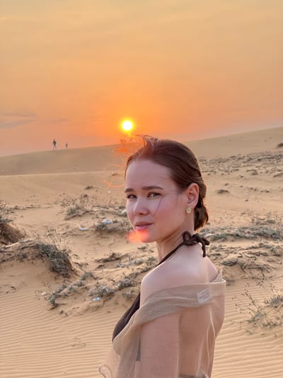 The image captures an adult female posing at sunset on sandy dunes near Phan Thiet, Vietnam. The subject, with brown hair pulled back and light skin, is positioned slightly off-center to the right, facing slightly left and looking directly at the camera. She wears a dark brown halter-neck top and a sheer, light brown, shimmering shawl draped over her shoulders. A circular gold-toned earring adorns her visible right ear. A prominent orange lens flare partially obscures her left cheek.
The background consists of extensive, undulating sand dunes with distinct ripple patterns. Sparse, dry vegetation, appearing as small bushes or grass clumps, is scattered across the dunes in the foreground and mid-ground. On a distant dune to the left, two silhouetted figures, one appearing adult and the other child-like, are visible. The sky exhibits a gradient of orange and yellow hues, with a bright, circular, yellow-orange sun low on the horizon, slightly right of the frame's center. Minor scattered light-colored debris is discernible among the vegetation in the mid-ground.