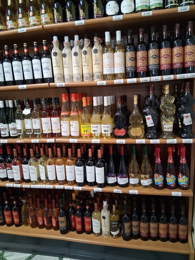 This is a photograph of a wall of wine bottles displayed in a store or cellar. The bottles are of various shapes and sizes, and most are dark glass with labels indicating different wines. The shelves are made of wood, and there are price tags visible on some of the bottles. The lighting is somewhat dim, suggesting an indoor setting.

The primary subjects are the wine bottles, arranged in neat rows. The labels are predominantly in Italian, with text such as "CRICOVA," "LIDIA," "PASTORAL," "CHARDONNAY," and "MERLOT." Some labels also include prices in Euros, such as "5.00€," "8.50€," and "3.99€." The arrangement suggests a retail environment, possibly a wine shop or a section of a larger store.

There are no people or animals visible in the image. The scene is static, with no apparent activity taking place other than the display of the products. The setting appears to be a commercial establishment, given the organized presentation of goods and pricing information. The overall impression is one of a well-stocked wine collection. The context provided indicates the location is Padua, Italy, which aligns with the Italian language visible on the labels.