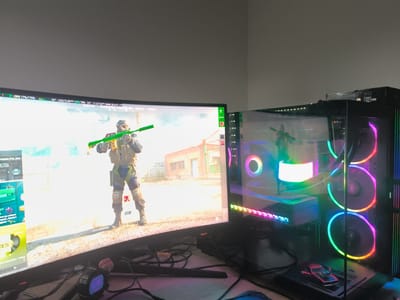 The image depicts a computer setup with a curved monitor displaying a first-person shooter game. A character with a gas mask and weapon is visible on the screen. The monitor also displays CPU and GPU usage statistics. To the right of the monitor is a desktop computer case with transparent panels and RGB lighting on the fans and components. A toy car is placed on top of the computer case. A smartwatch showing "15:55" is visible on the desk in front of the monitor. The background is a plain wall. There is no indication that this image is from Havixbeck, Germany.