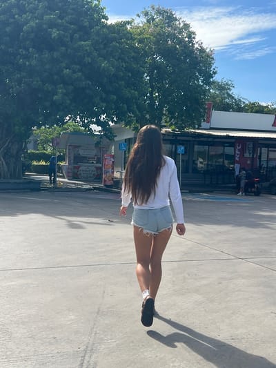 A young woman with long, dark hair stands in front of a KFC restaurant in Ban Ko Pru, Thailand. She is wearing a white long-sleeved shirt and light blue denim shorts. Her back is to the camera, and she is looking towards the KFC. The sun is shining, casting a long shadow on the pavement. Trees are visible to the left of the KFC, and a clear blue sky with white clouds is above. In the background, there are other buildings and some signage. One sign shows a price of "50". The scene appears to be during the daytime.