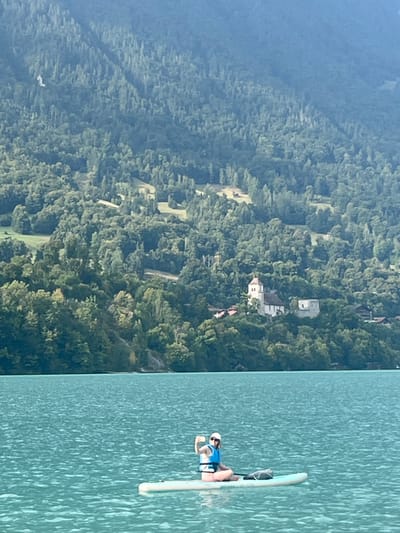 The scene depicts a body of water in the vicinity of Bönigen, Switzerland, with a forested mountain range in the background. In the mid-foreground, an individual is seated cross-legged on a light-colored stand-up paddleboard. The individual wears a white hat, sunglasses, a blue top, and a light blue personal flotation device. Their left arm is raised, with the hand clenched into a fist. A dark object is positioned on the paddleboard in front of the individual. The water exhibits a distinct turquoise hue with minimal surface ripples. Across the water, the lower slopes of a mountain are heavily forested. On a cleared section of the lower hillside, a white building with a prominent brown-capped bell tower is visible, adjacent to an older, light-colored stone structure, indicative of a church and an associated historical building. Further up the slopes, additional forested areas and small clearings with minor structures are discernible. A small, white avian object is present against the upper left section of the mountain.