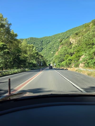The image shows a view from inside a vehicle driving on a paved road surrounded by lush green mountains and trees. The road is divided into two lanes with white and red markings. In the distance, a dark-colored vehicle is visible on the road. The sky is clear and blue. The scene appears to be a scenic drive through a mountainous region.
