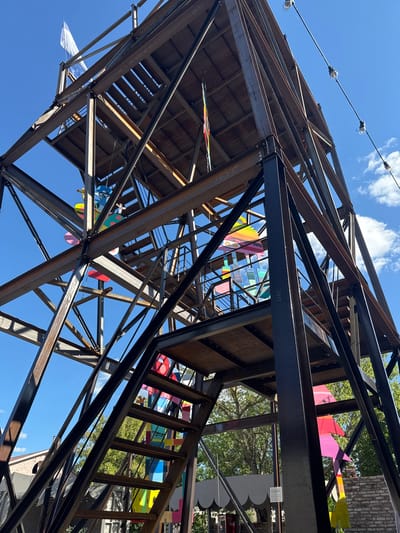 The image shows a large, industrial-style metal tower structure, likely an art installation or a part of an outdoor event space. The tower is made of steel beams and plates, with visible staircases and platforms. It is decorated with numerous brightly colored, abstract shapes and figures, resembling kites or hanging sculptures. A white flag is visible at the top of the structure. The sky is clear blue with some scattered white clouds. In the background, there are trees, and parts of buildings are visible, suggesting an urban or semi-urban outdoor environment. The overall impression is one of an artistic and possibly temporary structure.
The location is the city of Yelets, Russia. Specifically, the structure is located at the Yelets Museum of Local Lore.