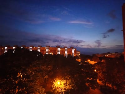 This is a nighttime, eye-level, medium-shot photograph of apartment buildings and trees in the city of Most, Czechia. The sky is a deep blue with scattered clouds. The apartment buildings, lit by numerous windows, are in the background. In the foreground, dark trees with some illuminated leaves suggest the presence of streetlights. The overall impression is one of a residential area at dusk or after dark.