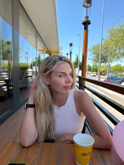 A blonde woman is seated at a wooden table outdoors, in front of a building with large glass windows, likely a McDonald's restaurant based on the McCafe cup. She is looking towards the camera with her right hand near her hair. She is wearing a white tank top and a watch on her left wrist. A pink balloon is partially visible to her right. The background shows a street with cars and trees under a clear blue sky. The location is identified as Húmera-somosaguas-prado Del Rey, Spain.