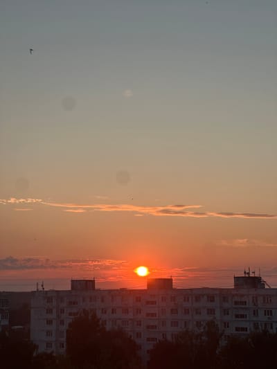 The image depicts a sunset over a city skyline, likely in Bakhchysarai, Russia. The sun, a bright orange orb, is positioned just above the horizon, casting a warm glow across the sky. A few scattered clouds, tinged with orange and pink, drift lazily above the buildings. The foreground is dominated by a row of multi-story apartment buildings, their facades weathered and utilitarian. Silhouetted trees stand in front of the buildings, their dark forms contrasting with the vibrant sky. Several small birds are visible in flight against the colorful backdrop. The overall impression is one of a tranquil evening scene in an urban environment.