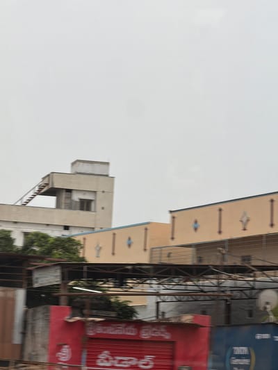 The image shows a street scene in Bethamcherla, India. In the background, there are several multi-story buildings. The building on the left is beige and has an external staircase leading to a rooftop area. It features several windows and a section with horizontal blinds. Adjacent to it is another building with a light brown facade adorned with decorative motifs that appear to be stylized diamonds and vertical lines. In the foreground, there are various structures, including a red building with a corrugated metal shutter and white text in a non-Latin script. To the right of the red building is a blue structure with text that includes "TATA Genuine". Trees are visible between the foreground and background buildings. The sky is overcast.