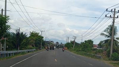 The image shows a two-lane asphalt road in Blangtambue, Indonesia. The road is lined with trees and vegetation on both sides. Several power lines are strung across the sky. There are a few vehicles on the road, including a truck, a white van, and two motorcycles. One motorcycle is in the foreground, ridden by a person wearing a red shirt and a helmet. The other motorcycle is further down the road, with a rider wearing a blue shirt and a helmet. The truck is in the middle distance, and the van is behind it. There are buildings visible in the background, as well as mountains. The sky is cloudy.