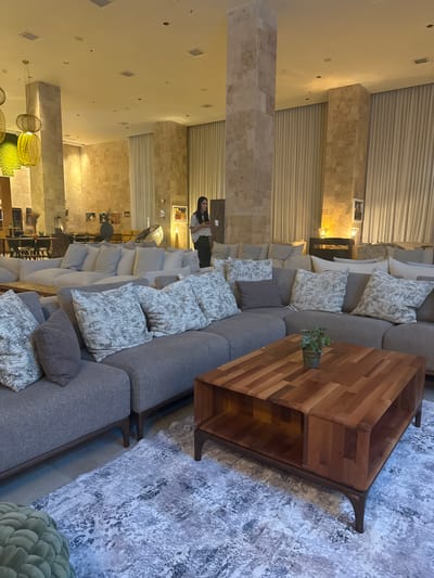 A woman stands holding a white cup in a well-lit hotel lobby in Tirat Karmel, Israel, with ample seating. A large, L-shaped gray sectional sofa dominates the foreground, adorned with several patterned throw pillows. In front of the sofa sits a rectangular wooden coffee table with an open base and a small potted plant on top. Behind the sectional, a lighter-colored modular sofa arrangement extends, also featuring numerous throw pillows. The background reveals textured stone walls, tall pillars, and floor-to-ceiling drapes. Hanging pendant lights with a woven texture are visible overhead, along with recessed lighting and ceiling speakers. Artwork is displayed on the walls. To the left, a dining area with tables and chairs is partially visible. The floor is covered with a light-colored patterned rug.