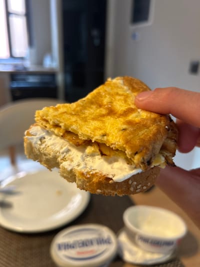 The scene depicts an indoor setting, likely a kitchen or dining area, located in the city of Archanes Municipal Unit, Greece. A person's right hand holds a partially consumed sandwich. The sandwich is constructed with a bottom layer of bread, possibly a baguette slice, featuring visible sesame seeds on its crust. This bread is liberally spread with a white, creamy substance, identified as cream cheese. Layered atop the cream cheese are several pieces of translucent, golden-brown caramelized onions. The topmost layer is a folded, golden-yellow omelet, exhibiting slight browning and dark specks, possibly herbs. A bite has been taken from the front-left portion of the sandwich. In the blurred mid-ground, on a dark, textured surface, a white dinner plate and a metallic utensil (fork or knife) are visible to the left. To the right, two white plastic containers of "Philadelphia" brand cream cheese are present; one is closed and clear, while the other is open and partially utilized. The background is blurred, showing a window with natural light entering from the left and dark furniture or appliances on the right.