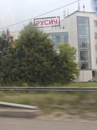 The image shows a modern, multi-story building with large windows, likely a commercial or office space. A prominent red sign with white lettering is visible on the facade, reading "РУСИЧ" (Rusich). Below this, a smaller sign with Russian text is present. Several trees are in the foreground, partially obscuring the lower levels of the building. A metal guardrail runs horizontally across the middle ground. In the background, a car is parked near the building. The sky appears overcast. The overall impression is a scene captured from a moving vehicle on a road adjacent to the building, possibly in Mikhalkovo, Russia, as suggested by the signage.