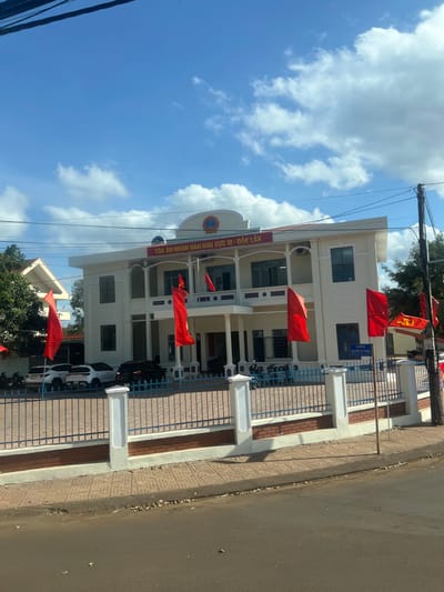 This image depicts the exterior of a two-story institutional building, identified by the banner "TÒA ÁN NHÂN DÂN KHU VỰC 10 - ĐẮK LẮK" (People's Court of Area 10 - Đắk Lắk), located in Trung Hồ, Vietnam. The building is light cream or off-white with a central entrance featuring columns supporting an upper balcony, above which a judicial emblem of scales is visible. Numerous rectangular windows are set into the facade.
In front of the building, multiple red flags on poles are displayed; one on the far right includes the yellow hammer and sickle emblem. Behind a blue metal fence with white concrete pillars and a brick-patterned base, several vehicles are parked: two white SUVs, one dark sedan, and multiple motorcycles/scooters.
The scene is under a clear blue sky with scattered white cumulus clouds. In the immediate foreground, a paved sidewalk with a geometric pattern separates an asphalt road from the fence. A utility pole with power lines is visible on the right side of the frame, alongside a partially visible blue street sign bearing Vietnamese text, likely "ĐƯỜNG NGUYỄN TẤT THÀNH." Green foliage and parts of another building are present in the background to the left. No individuals are clearly discernible.