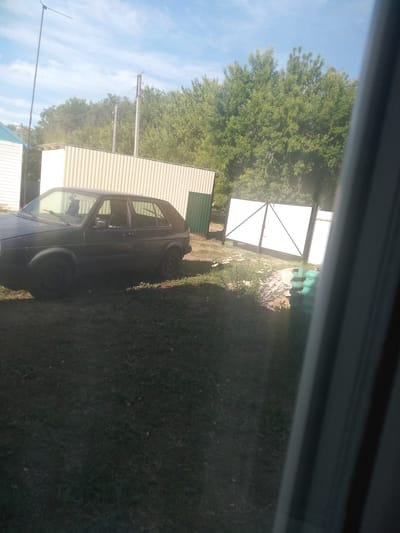 The image depicts a dark-colored, older model hatchback car parked on a grassy area. The car is positioned to the left of a corrugated metal structure. In the background, there is a white gate with a black frame and a green fence. Beyond the gate and fence are lush green trees, indicative of an outdoor, possibly rural or suburban, setting. The sky above is blue with scattered white clouds. The scene appears to be an exterior shot, possibly in a yard or open space. The area is well-lit by natural sunlight. There are no people visible in the image. The overall impression is of a still, daytime scene.