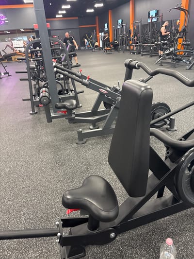 The image, rotated 90 degrees counter-clockwise, captures the interior of a modern gym or fitness center in Trilistnik, Bulgaria. The flooring consists of a grey speckled rubber mat, while the walls are a mix of dark grey/black and bright orange panels. Bright artificial overhead lights illuminate the space.
In the immediate foreground, a large, black-framed piece of gym equipment, likely an upper body machine, is prominent. It features black padded rests, including a rectangular pad and a bicycle-style seat with a visible red adjustment lever. A clear plastic water bottle with a pink cap sits on the floor nearby.
The background showcases numerous other pieces of gym equipment, including various weight machines, benches, and racks, maintaining a consistent aesthetic with black frames and some orange details. Several monitors are mounted on poles throughout the facility. The brand name "PANATTA" is visible on one piece of equipment in the mid-ground. Faintly, outdoor scenery, possibly buildings or trees, can be seen through a distant opening. A purple-lit sign in the lower left background displays partial text, including "RM SELF."
Several people are visible throughout the gym, engaged in exercise. A man with a bald head and beard, wearing a dark tank top, is actively using a weight machine in the middle ground, appearing to perform a seated row. Other individuals are dispersed, utilizing different machines, indicating a typical gym environment during an active training session.