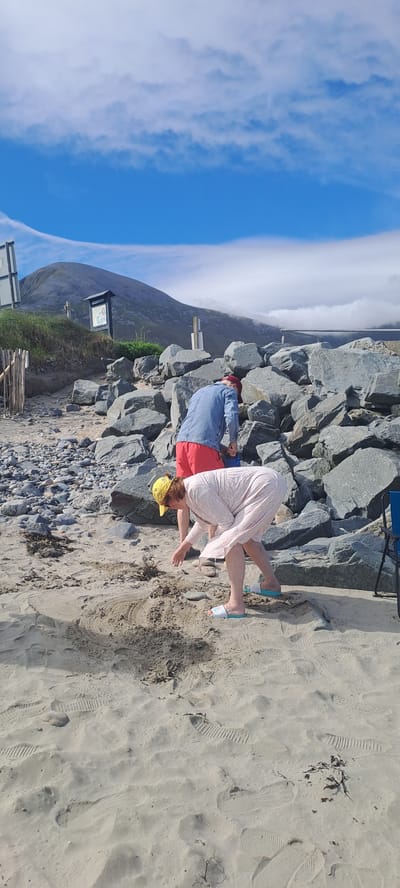Two individuals are present on a beach near a rocky coastline. The individual in the foreground is a woman wearing a pink dress and a yellow hat, bent over and appearing to interact with the sand. The second individual, a person in a denim jacket and red shorts, is standing behind her and also appears to be engaged in an activity with the sand. The environment consists of sand, rocks, and a glimpse of a hill and cloudy sky in the background, suggesting a coastal setting. The overall scene is one of casual outdoor activity.