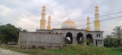 This is a horizontal, full-shot photograph of a mosque under construction in Buketglumpang, Indonesia. The mosque features a large, golden dome adorned with white diamond-shaped patterns and flanked by two tall, yellow minarets with white zigzag designs. In the foreground, a grey concrete wall with small rectangular openings runs across the width of the image. A concrete building with arched doorways is visible to the right of the wall and in front of the mosque's main structure. Palm trees and other greenery surround the mosque. The sky is overcast with a grey, cloudy appearance. There are power lines running horizontally across the image in front of the mosque and minarets. The bottom left corner displays a timestamp and a phone brand: "vivo V25 10 Jul 2026, 18:26". There are no people visible in the frame.